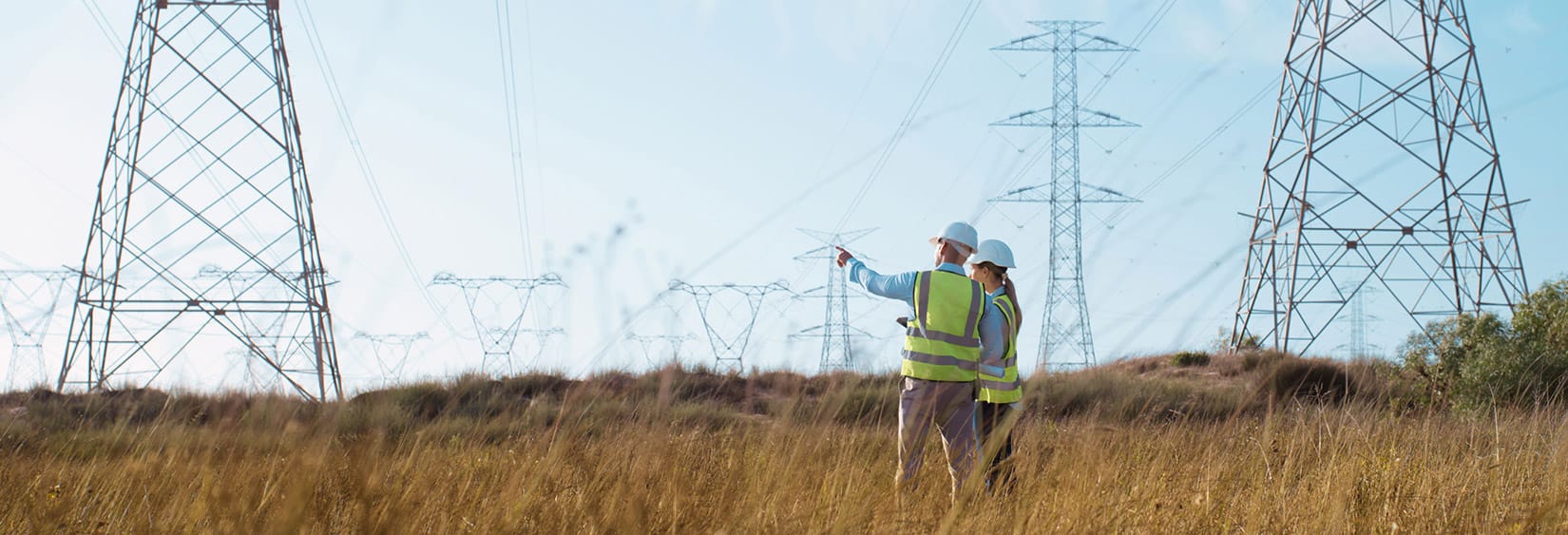 Two utility workers in safety vests and helmets inspect power transmission towers in a grassy field.